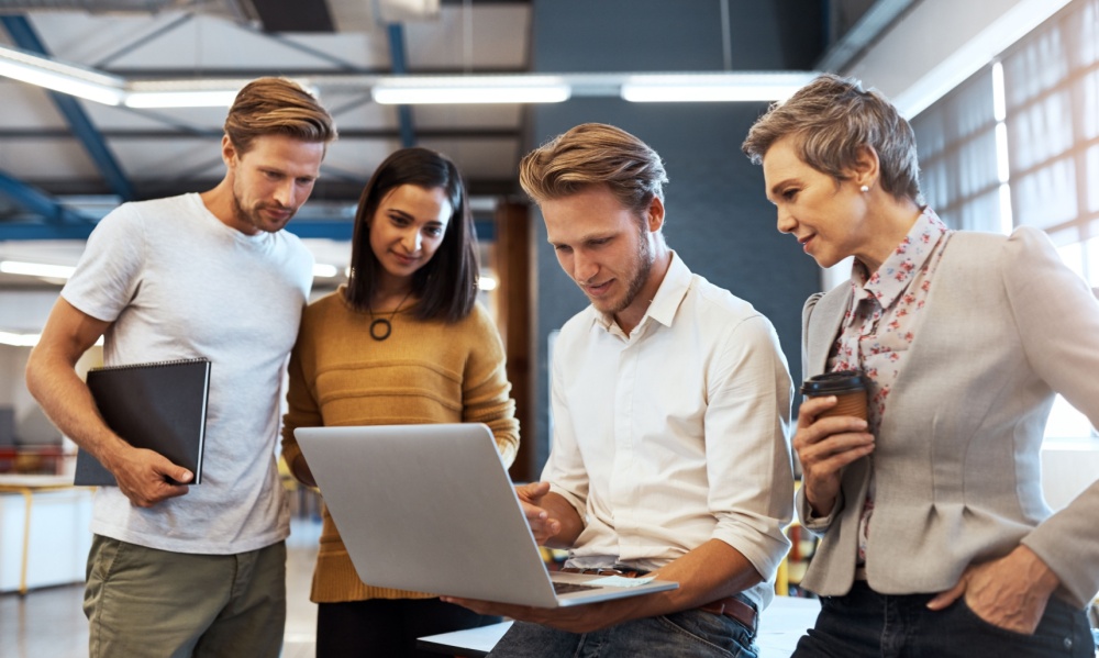 A man and two women look at a young blond man's computer during a startup meeting.